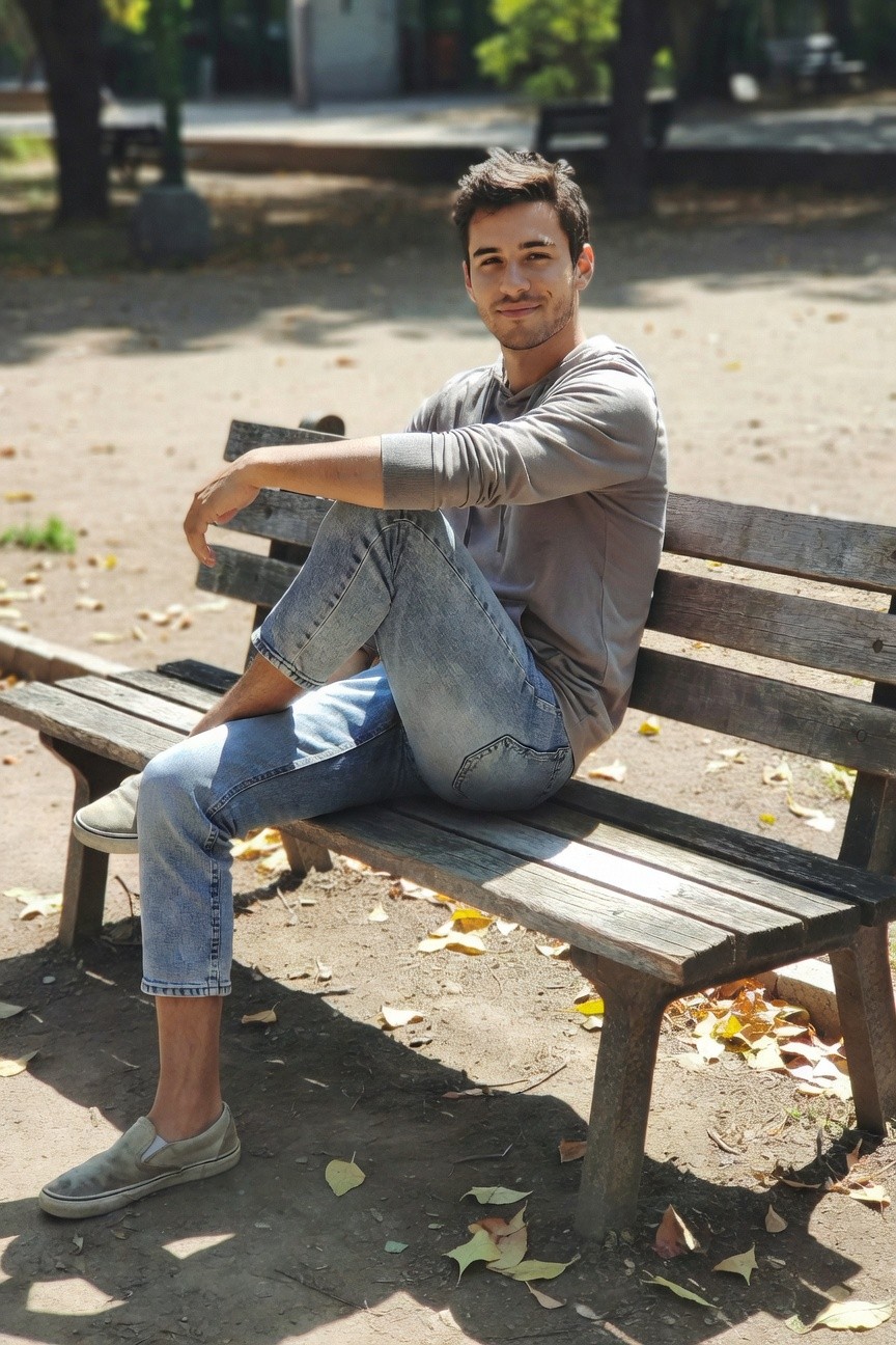 Man seated casually on wooden bench wearing light gray long-sleeve shirt, slim faded light blue jeans cuffed at ankles, and white slip-on sneakers