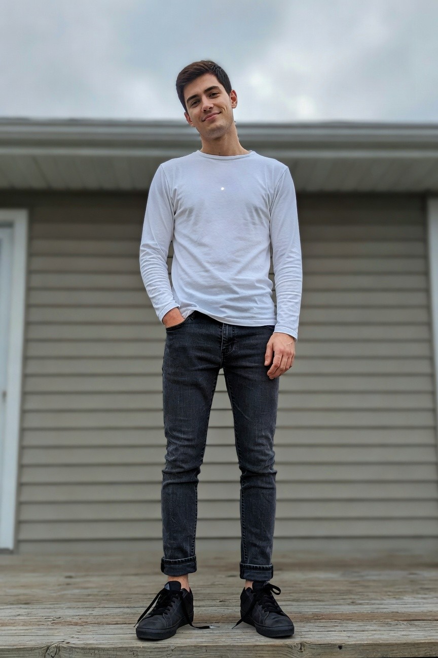 Young man in slim black cuffed jeans, white long-sleeve t-shirt, and black sneakers, standing casually with hands in pockets on a wooden deck