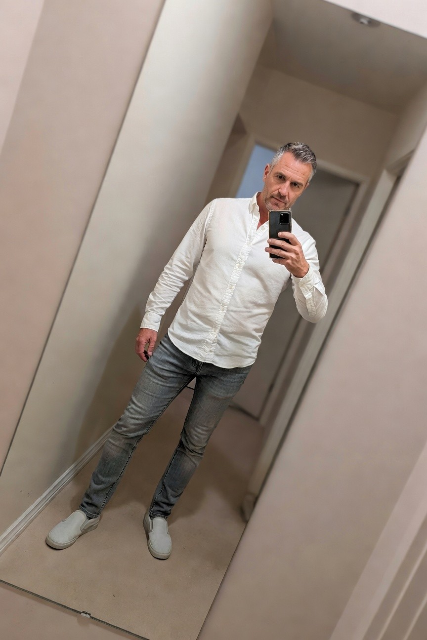 Man taking mirror selfie in white long-sleeve button-up shirt, gray distressed straight-leg jeans, and white slip-on sneakers, hallway setting