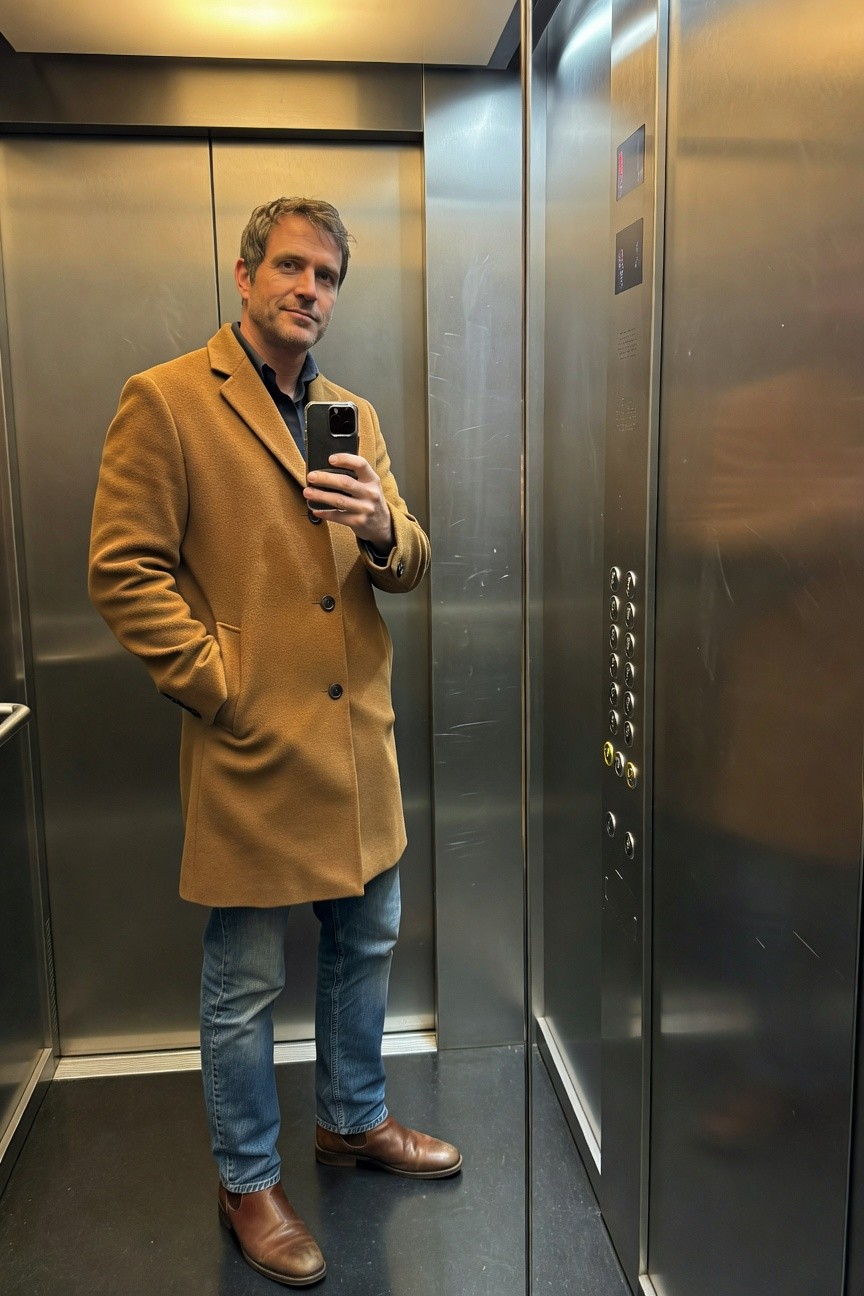 Man taking a mirror selfie in an elevator, wearing a long camel wool overcoat over a dark shirt, straight-leg faded blue jeans, and brown leather dress shoes, one hand holding phone and the other in pocket