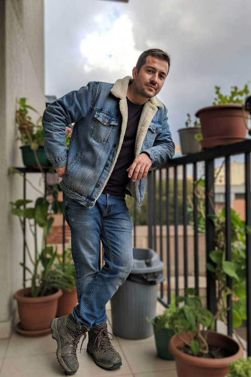 Man leaning casually on balcony railing in light wash shearling-trimmed denim jacket over dark crewneck sweater, straight blue jeans, and brown laced hiking boots, surrounded by potted plants
