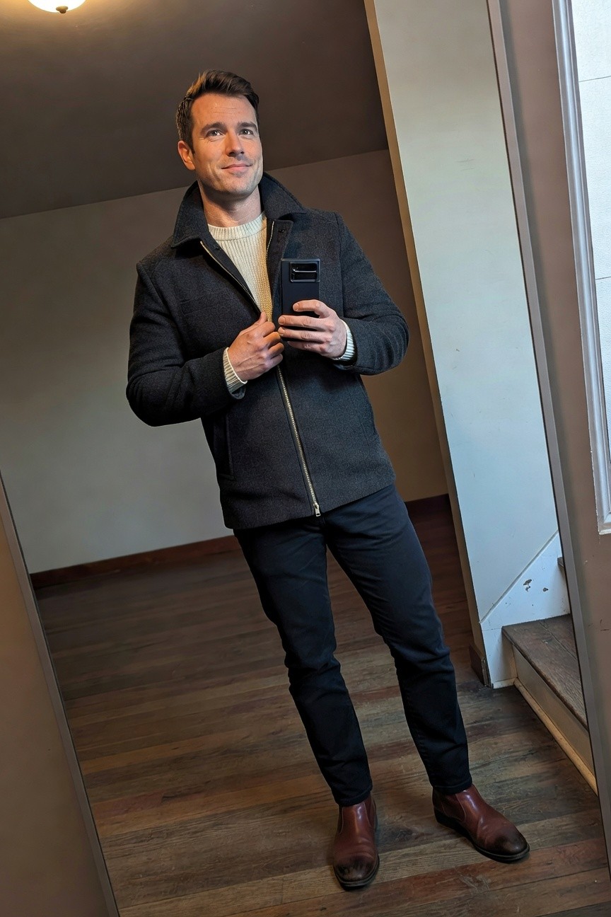 Man in dark grey wool jacket over cream turtleneck, black slim jeans, and brown leather Chelsea boots, posing in front of mirror indoors