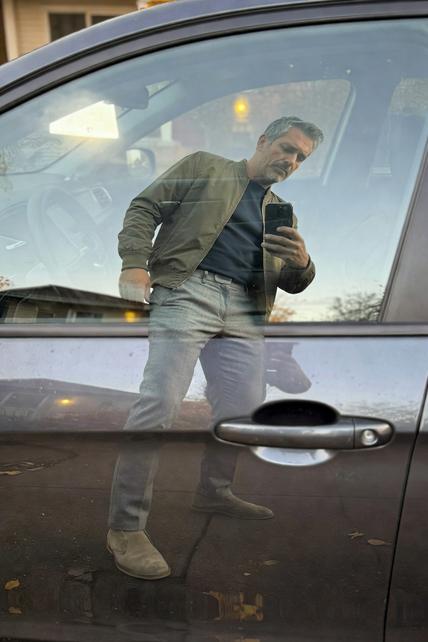 Man reflected in car window wearing olive green bomber jacket over navy t-shirt, light grey textured pants, and tan suede Chelsea boots, holding phone for selfie