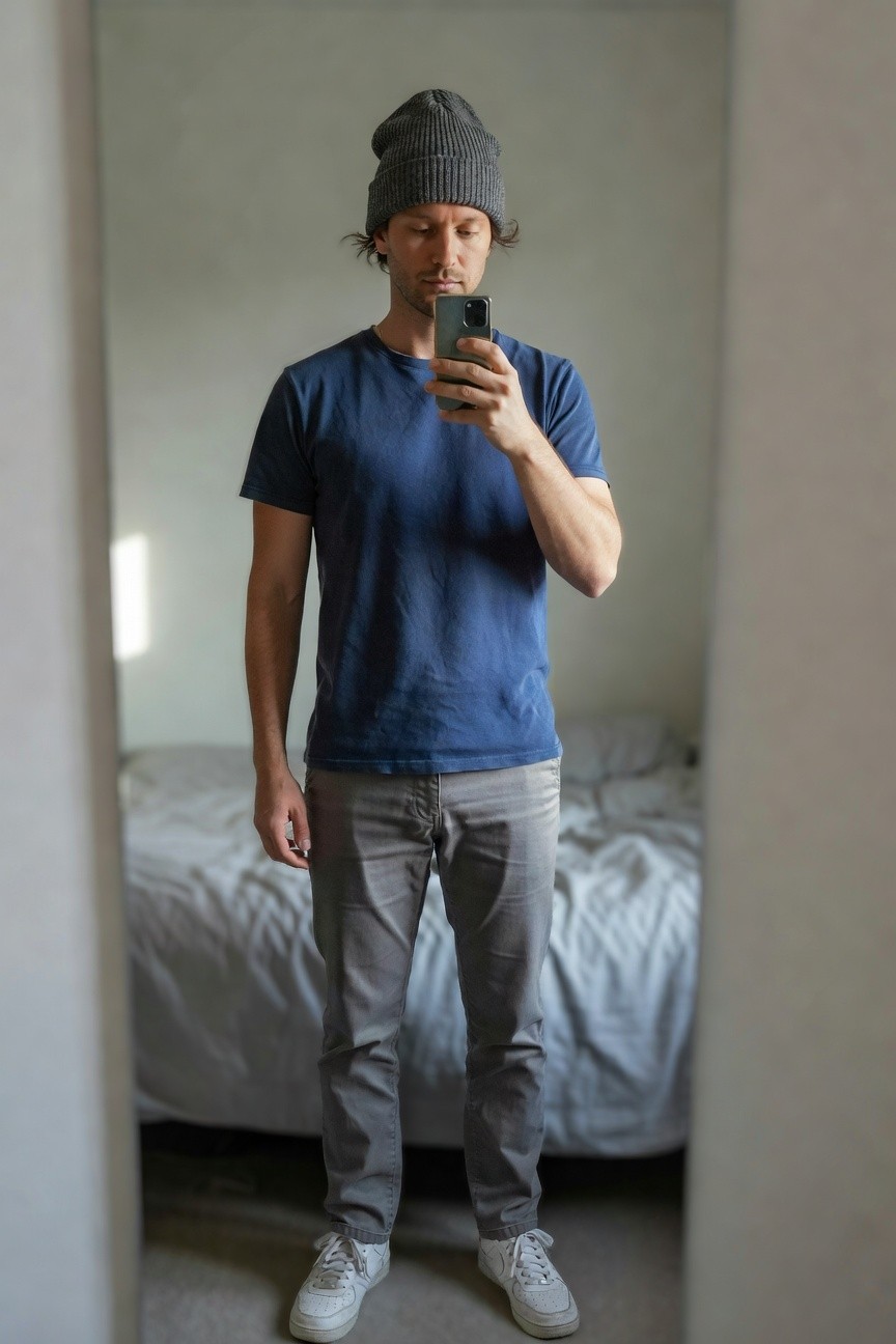 Man in a mirror selfie wearing a grey knit beanie, navy short-sleeve t-shirt, slim grey pants, white sneakers, and a thin chain necklace, standing in a simple bedroom