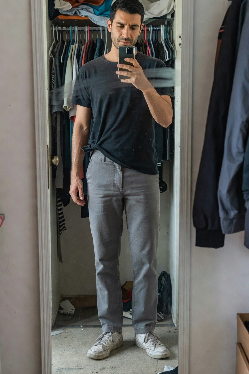 Man taking mirror selfie in open closet wearing loose black short-sleeve t-shirt tied at waist, straight-leg grey pants, and white sneakers, surrounded by hanging clothes and storage bins
