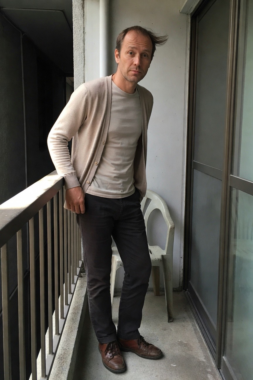 Man leaning on balcony railing in open light beige knit cardigan over white v-neck tee, slim dark grey pants, and brown leather boots, casual off-duty style