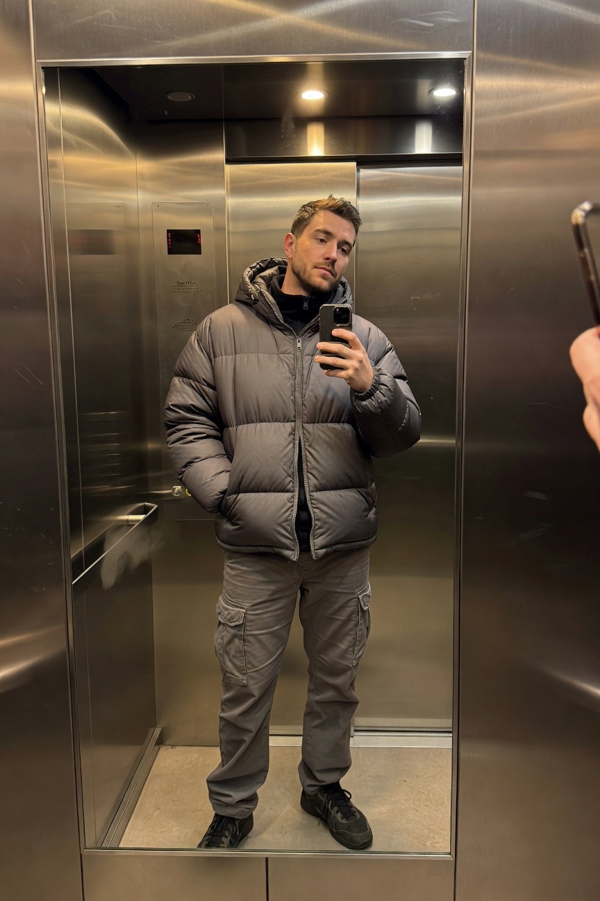 Man taking a mirror selfie in an elevator, wearing a baggy grey puffer jacket over black turtleneck, baggy grey cargo pants, and black chunky sneakers