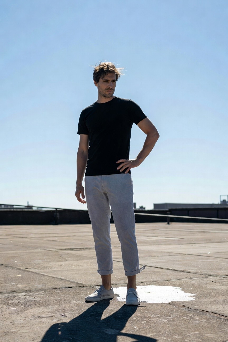 Man standing on rooftop wearing black short-sleeve t-shirt, light grey cropped chinos rolled at ankles, white lace-up sneakers, hand on hip, looking sideways against blue sky