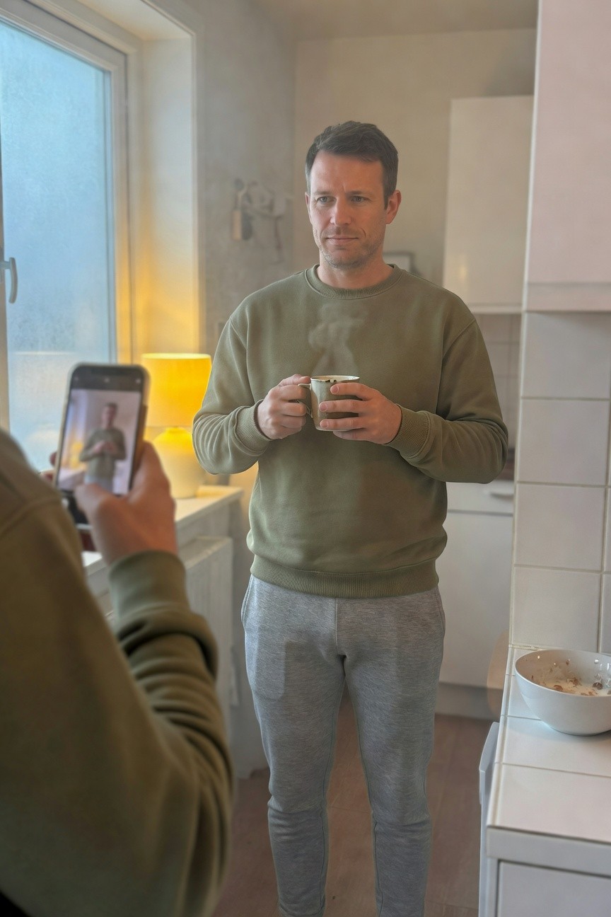 Man standing in kitchen wearing sage green crewneck sweatshirt and light grey joggers, holding two white mugs of coffee with phone screen showing his reflection, yellow lamp and white cabinets in background