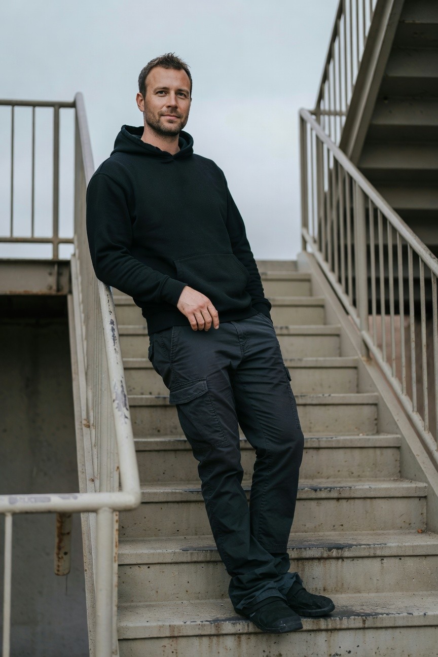 Man in black hoodie and dark grey cargo pants leaning against metal staircase railing, black sneakers on concrete steps