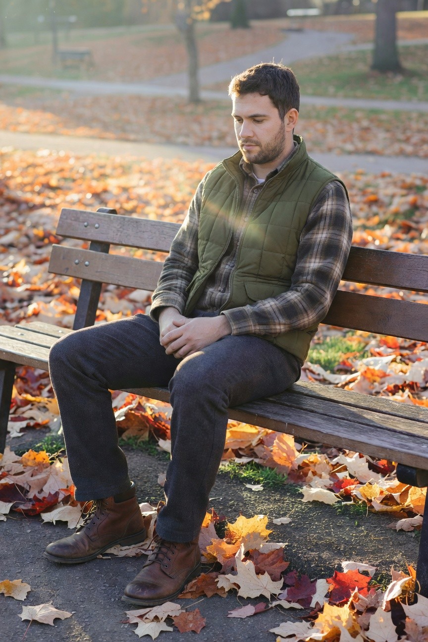 Man in dark grey slim pants, dark plaid flannel shirt, olive green quilted vest, and brown leather boots, sitting on wooden bench with hands clasped, autumn leaves on ground