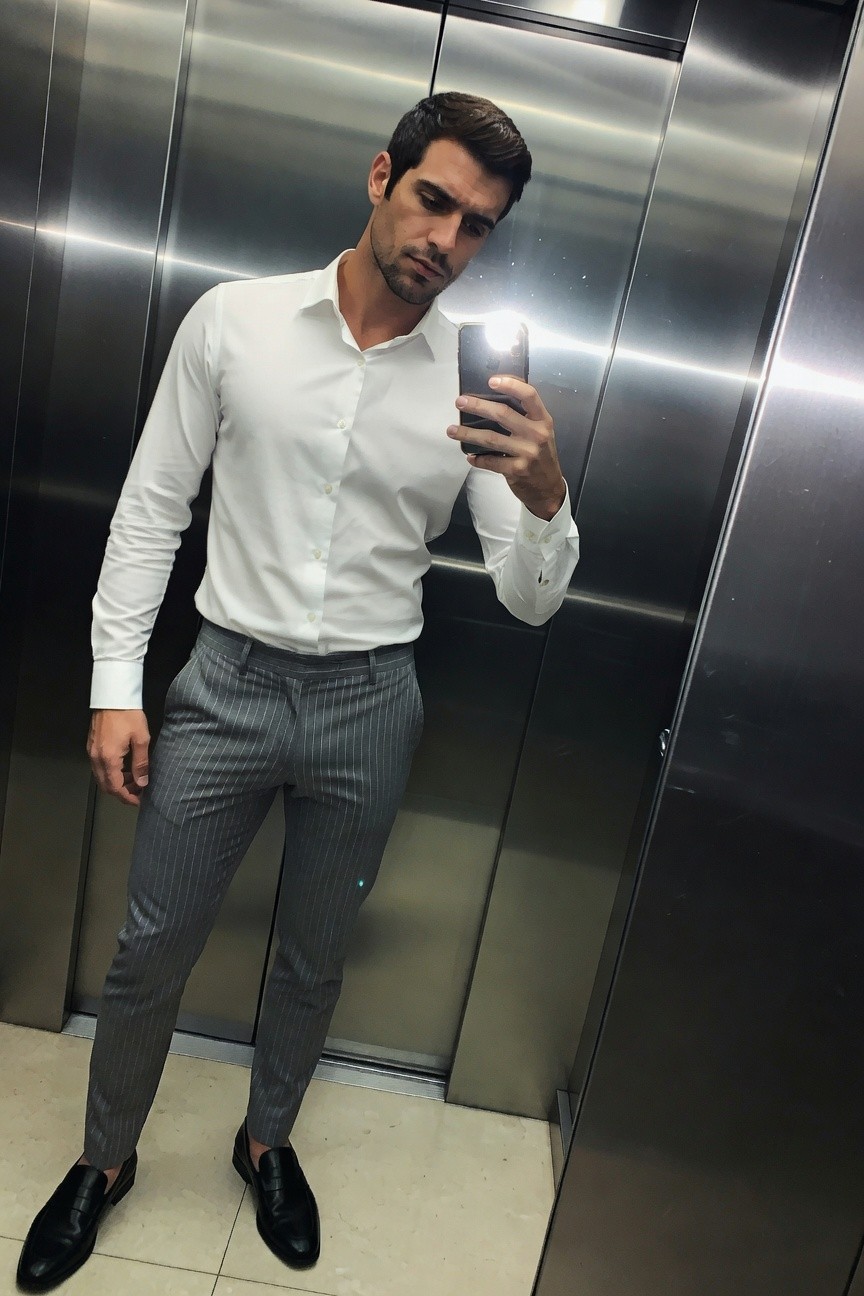 Man in elevator taking selfie, wearing white long-sleeve button-up shirt tucked into slim grey pinstripe trousers and black leather loafers