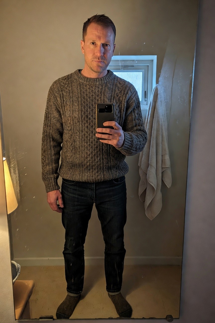Man taking mirror selfie in bathroom wearing gray-brown cable knit sweater, dark straight-leg jeans, and brown crew socks, holding phone