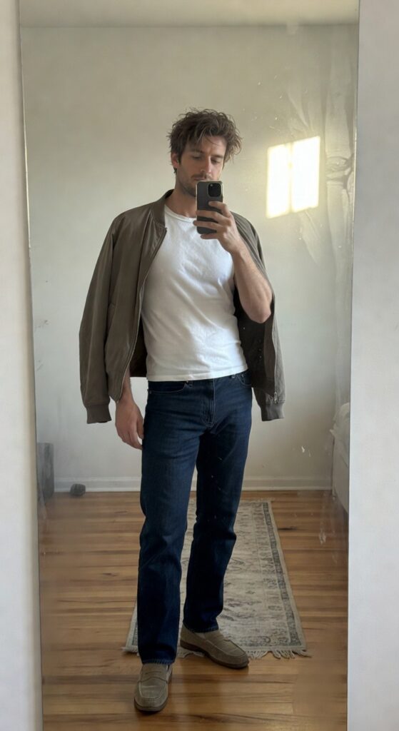 Mirror selfie of man wearing open tan suede bomber jacket over white t-shirt tucked into straight-leg dark blue jeans and tan suede loafers standing on wood floor with rug
