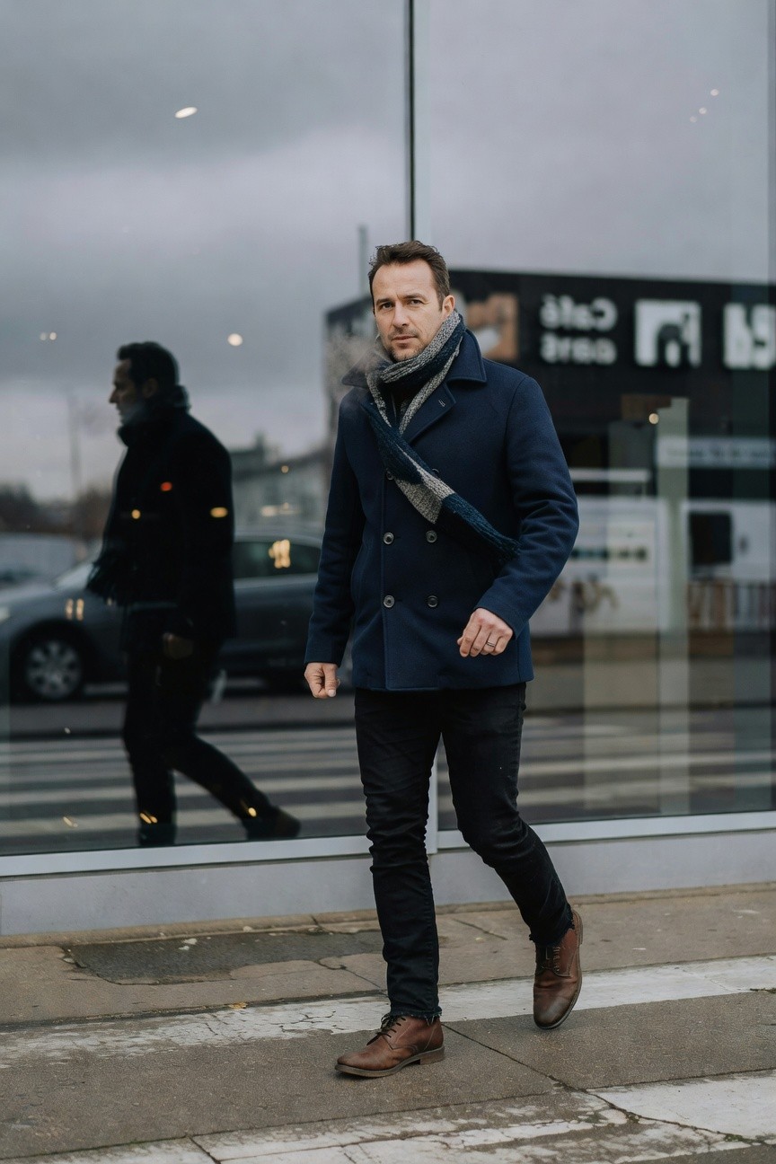 Man walking on crosswalk in navy double-breasted wool peacoat, gray-blue striped scarf, slim black jeans, and brown leather boots, reflections in glass building behind