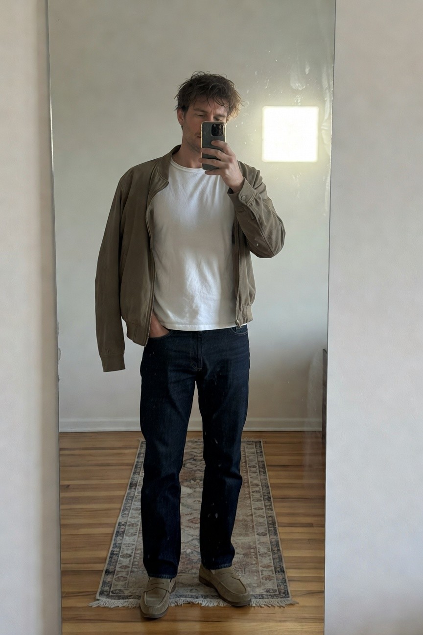 Mirror selfie of man wearing open tan suede bomber jacket over white t-shirt tucked into straight-leg dark blue jeans and tan suede loafers standing on wood floor with rug