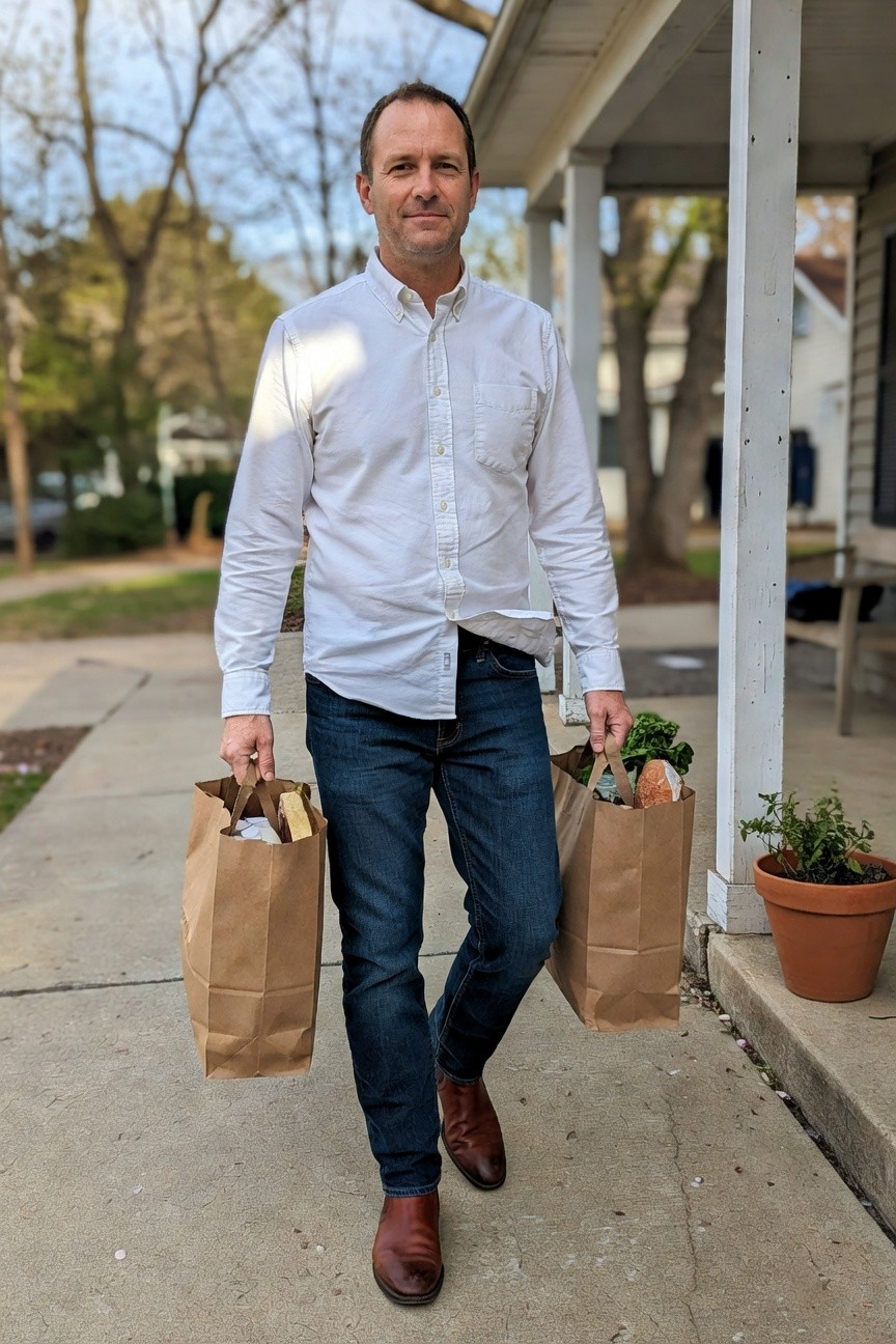 Man in white long-sleeve button-up shirt tucked into blue jeans, brown leather dress shoes, holding two brown grocery bags with bread and greens visible, standing on a sidewalk