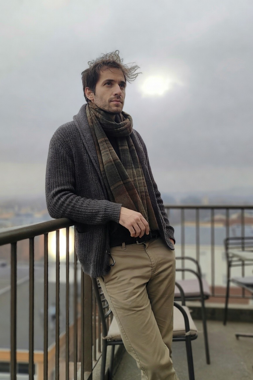 Man in open gray knit cardigan layered over plaid scarf in earthy tones, khaki chinos, and belt, leaning casually on a railing