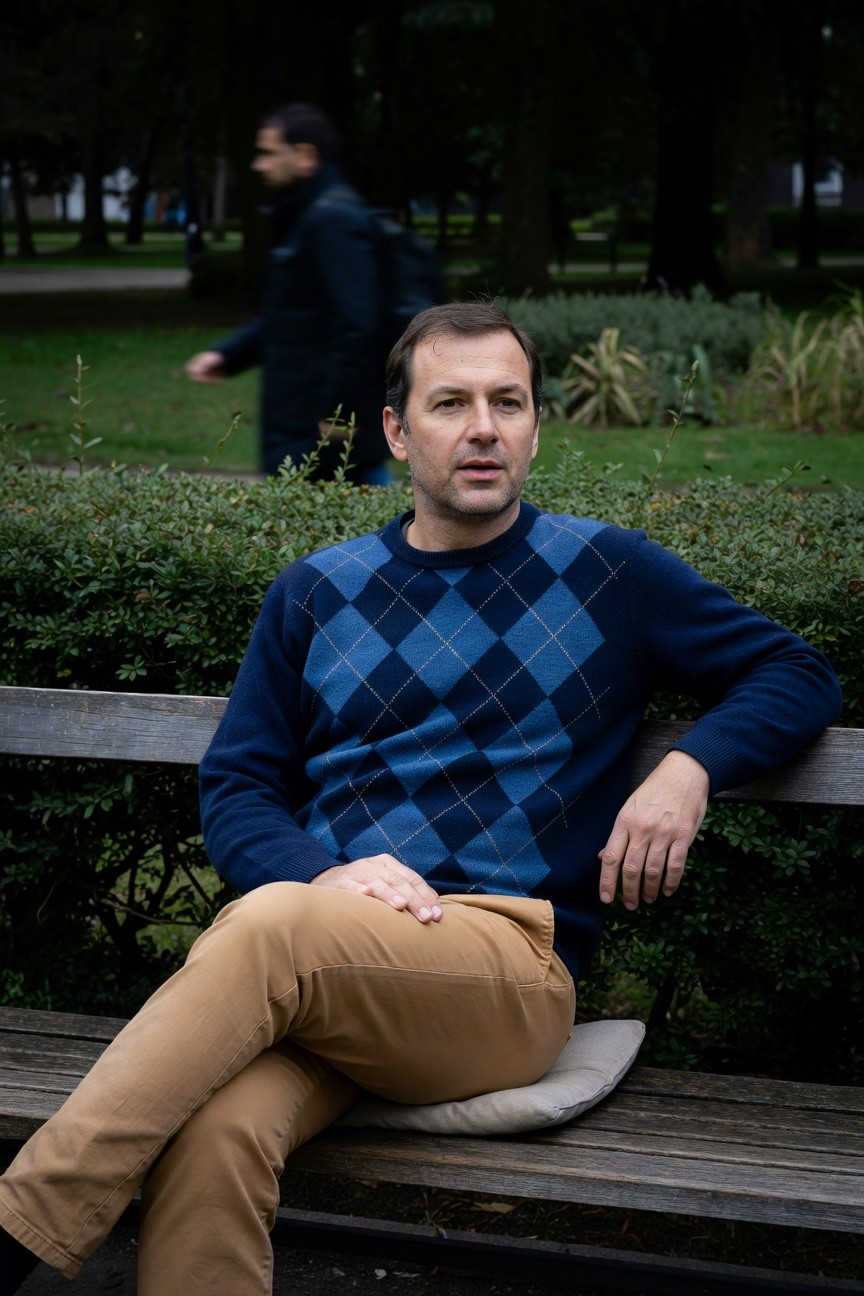 Man in his 40s sitting casually on a wooden park bench, wearing a navy blue argyle knit sweater with diamond pattern, beige chinos, and a light gray cushion under him, arm resting on bench back, green shrubs and park path blurred behind