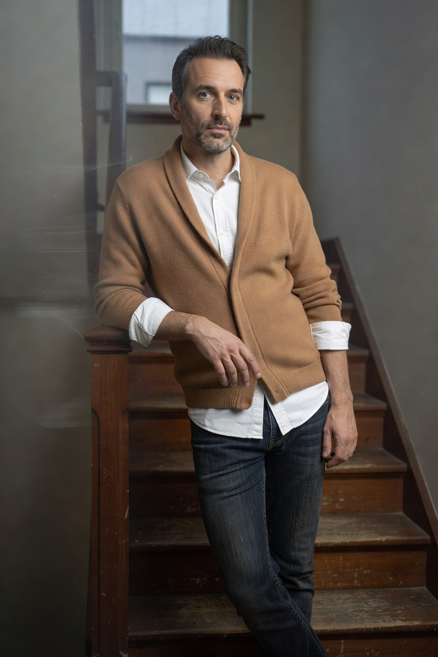 Handsome man in his 40s with short dark hair and goatee, wearing an open camel knit cardigan over a white button-up shirt with rolled cuffs, dark slim-fit jeans, leaning casually on a wooden staircase railing indoors