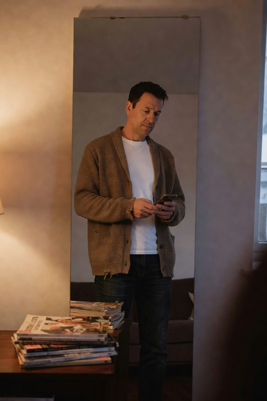 Man in open tan knit cardigan over white crewneck t-shirt and dark straight-leg jeans, holding phone while standing near a table with stacked magazines