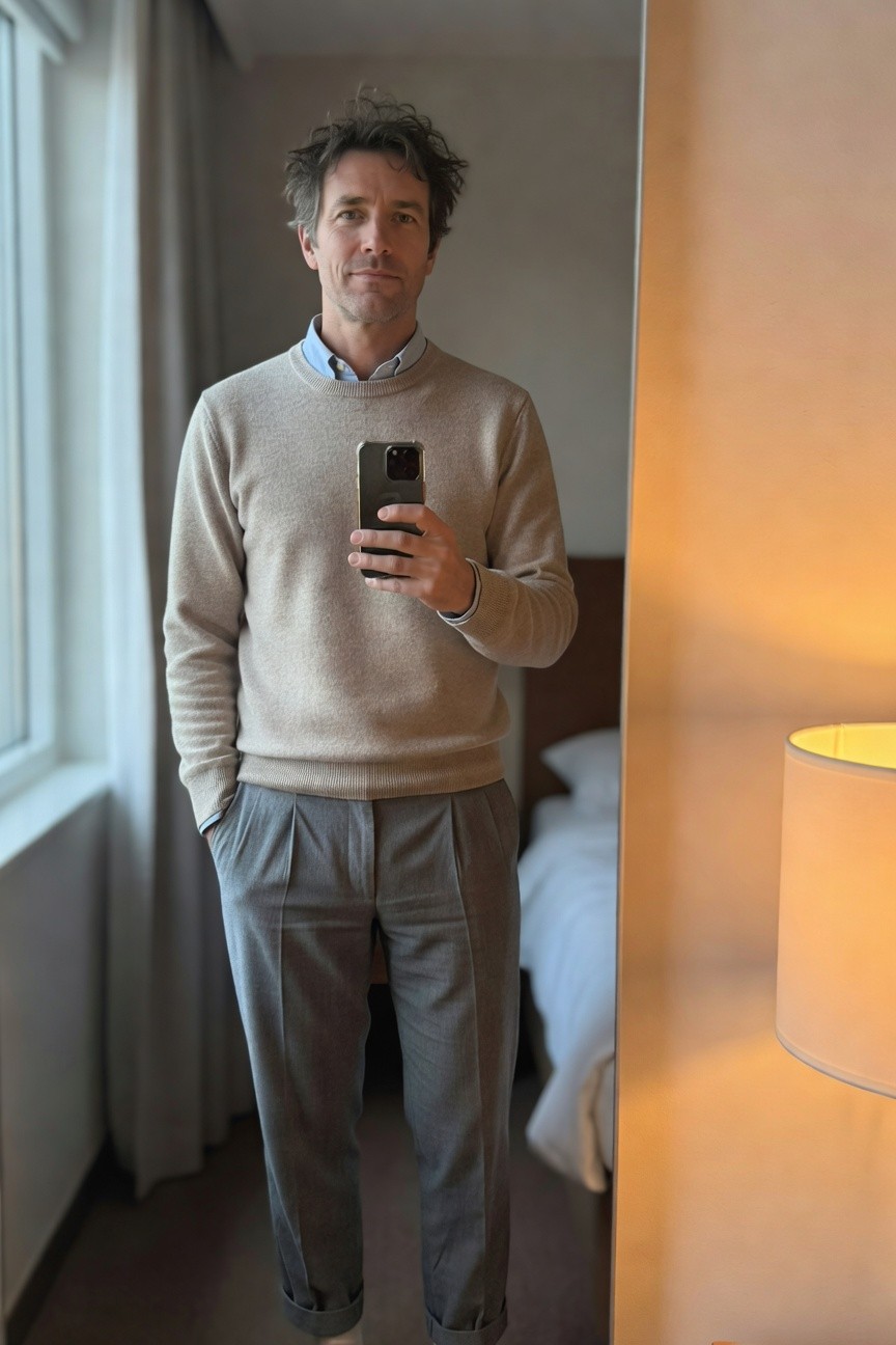Man in a light cream crewneck sweater over a pale blue collared shirt, paired with gray pleated trousers cuffed at the ankles, standing in front of a mirror with hands in pockets holding a phone, neutral hotel room background