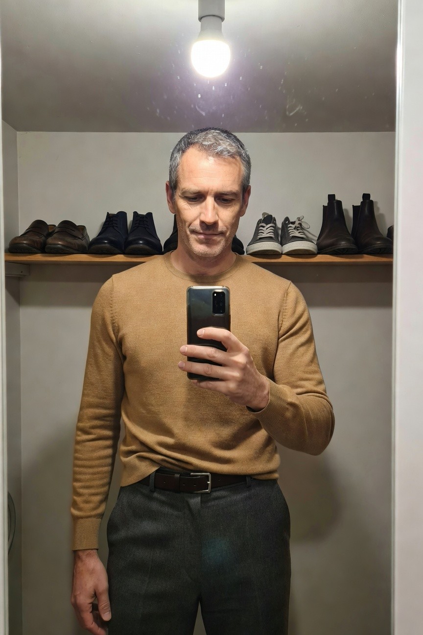 Middle-aged man with graying hair standing in a closet wearing a tan crewneck sweater, charcoal gray wool trousers, and brown belt, holding a smartphone for a selfie, with shoe shelves visible behind him