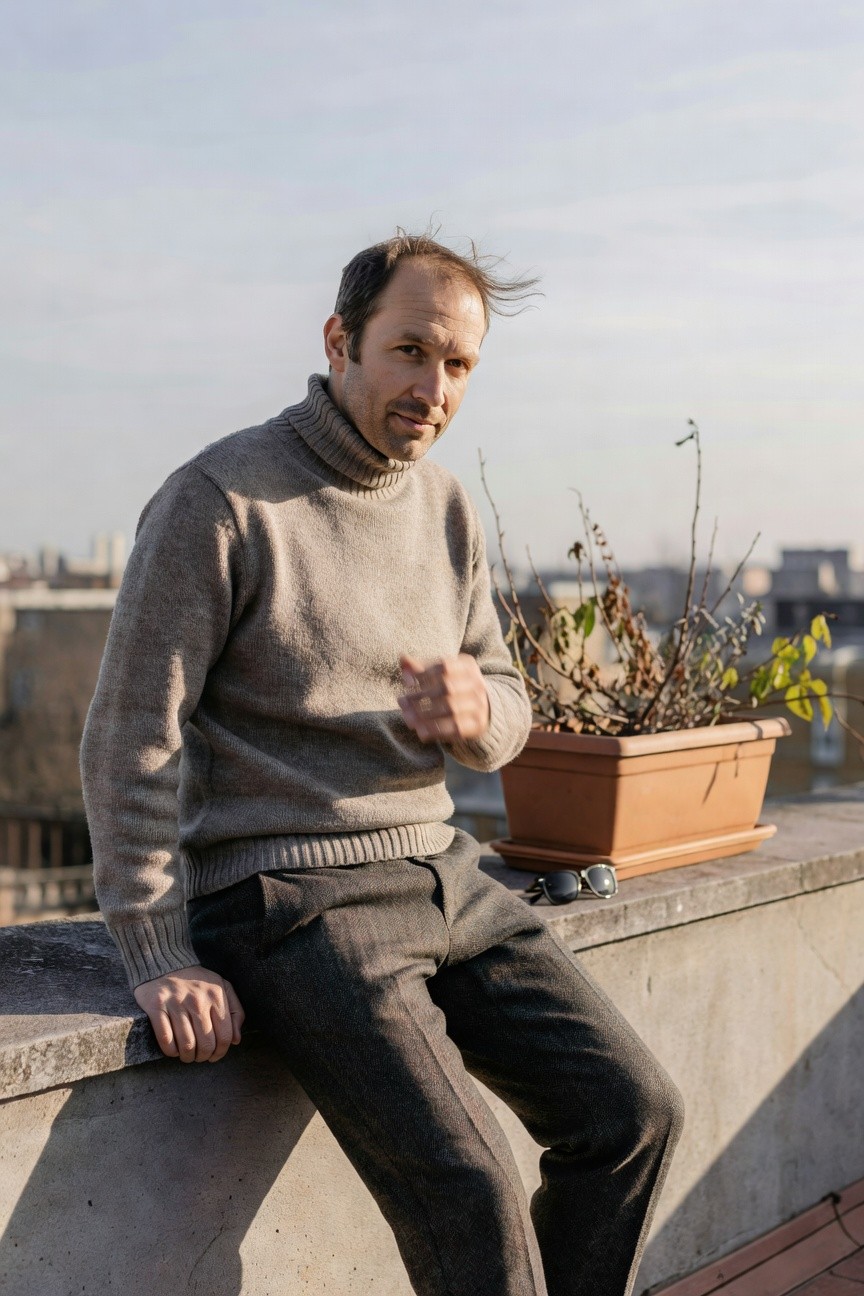 Man in pale gray turtleneck sweater and darker gray wool trousers sitting on concrete ledge with hand gesturing and sunglasses nearby