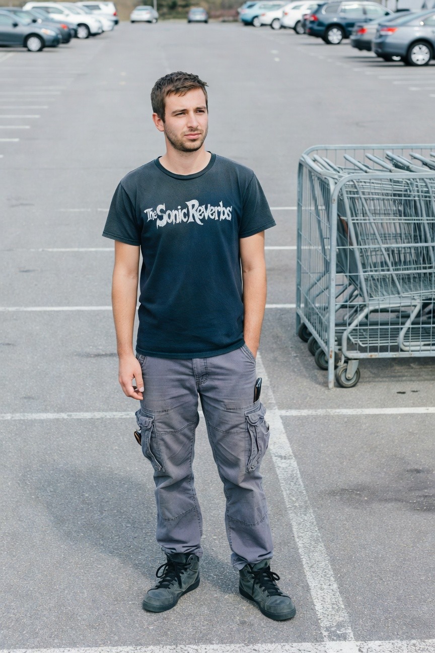 Young man in dark navy band t-shirt, grey cargo pants with multiple pockets, and black high-top sneakers standing casually near metal shopping cart racks in a parking lot