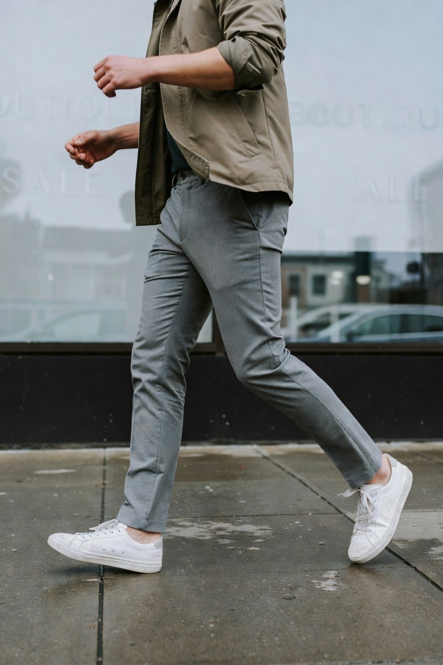 Man walking in slim grey pants, beige field jacket, dark tee, and white sneakers on wet urban sidewalk, side profile view emphasizing casual movement and neutral tones