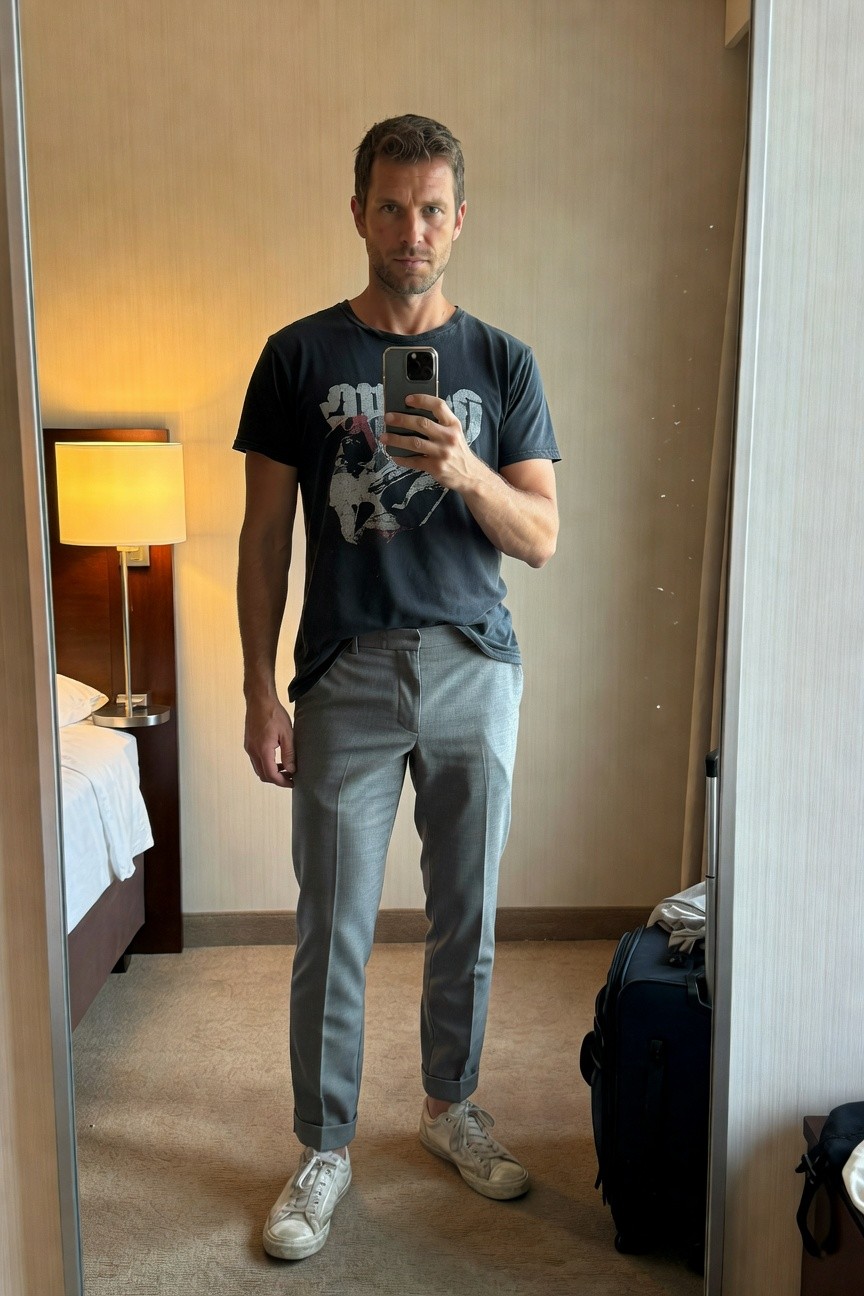 Mirror selfie of a man in a black graphic t-shirt with earth design, slim grey trousers cuffed at ankles, and white sneakers, standing in a hotel room with suitcase nearby