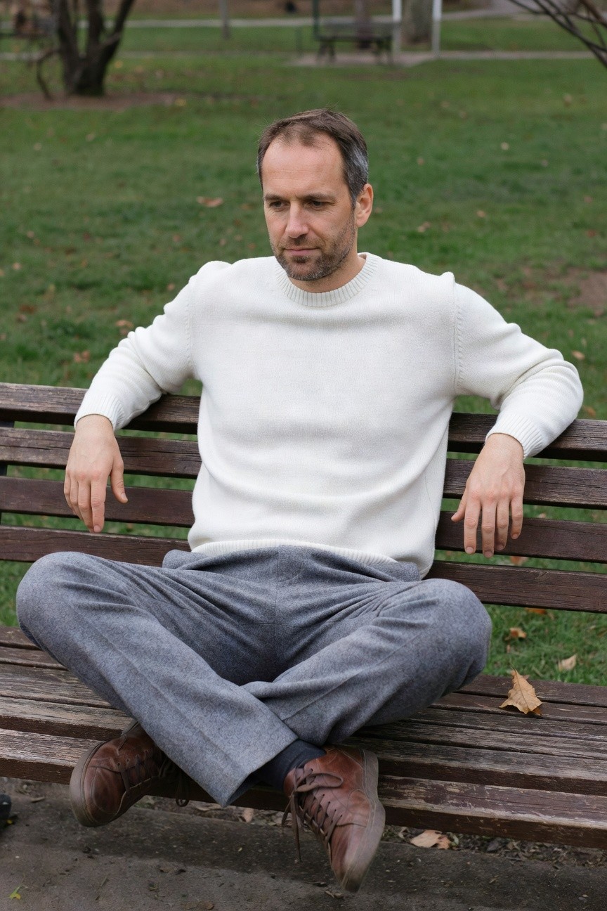 Man in white crewneck sweater, grey wool trousers, and brown leather shoes sitting cross-legged on a wooden bench in a park