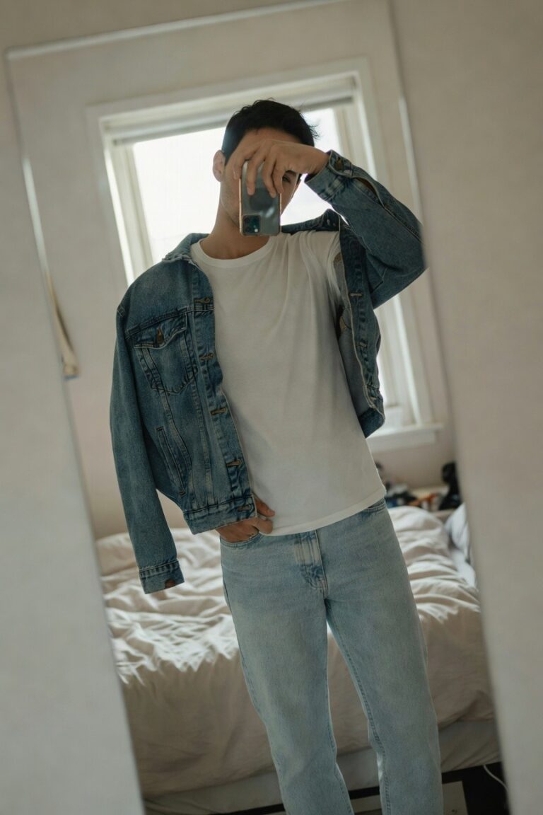 Man in mirror selfie wearing open medium-wash denim jacket over white crewneck t-shirt, light blue straight-leg jeans, silver watch on left wrist, bedroom setting with bed and clothes in background