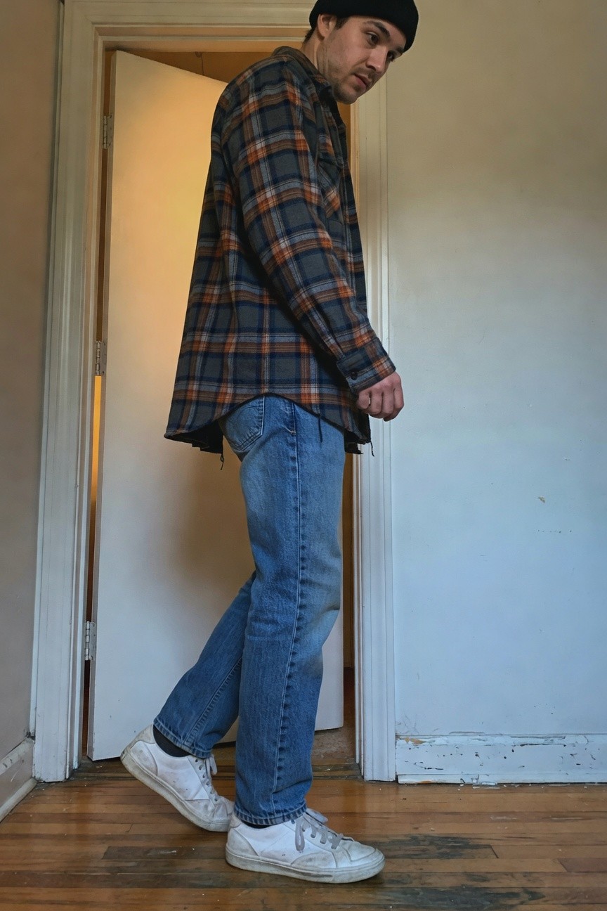 Side view of a man in a loose navy plaid flannel shirt untucked over light blue straight-leg jeans, white low-top sneakers, and black beanie, standing casually by an open interior door on hardwood floor