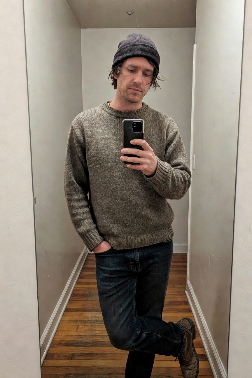 Man in mirror selfie wearing chunky mottled gray knit crewneck sweater, matching gray beanie, dark straight-leg jeans, and brown leather boots, casual pose with one hand in pocket.