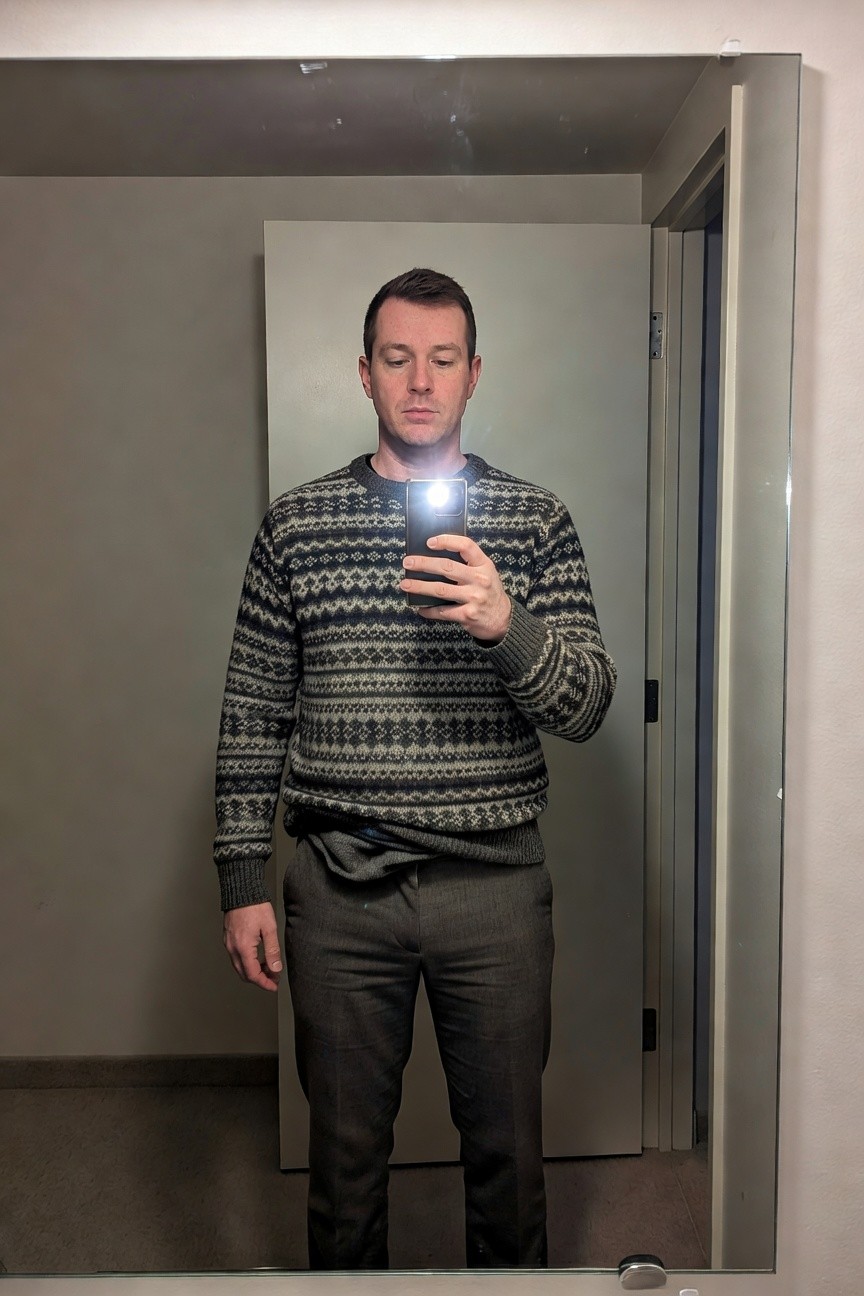 Man in a mirror selfie wearing a grey Fair Isle knit sweater with black and white Nordic patterns tucked into slim grey wool trousers with a belt, phone flash illuminating the outfit in a plain indoor room