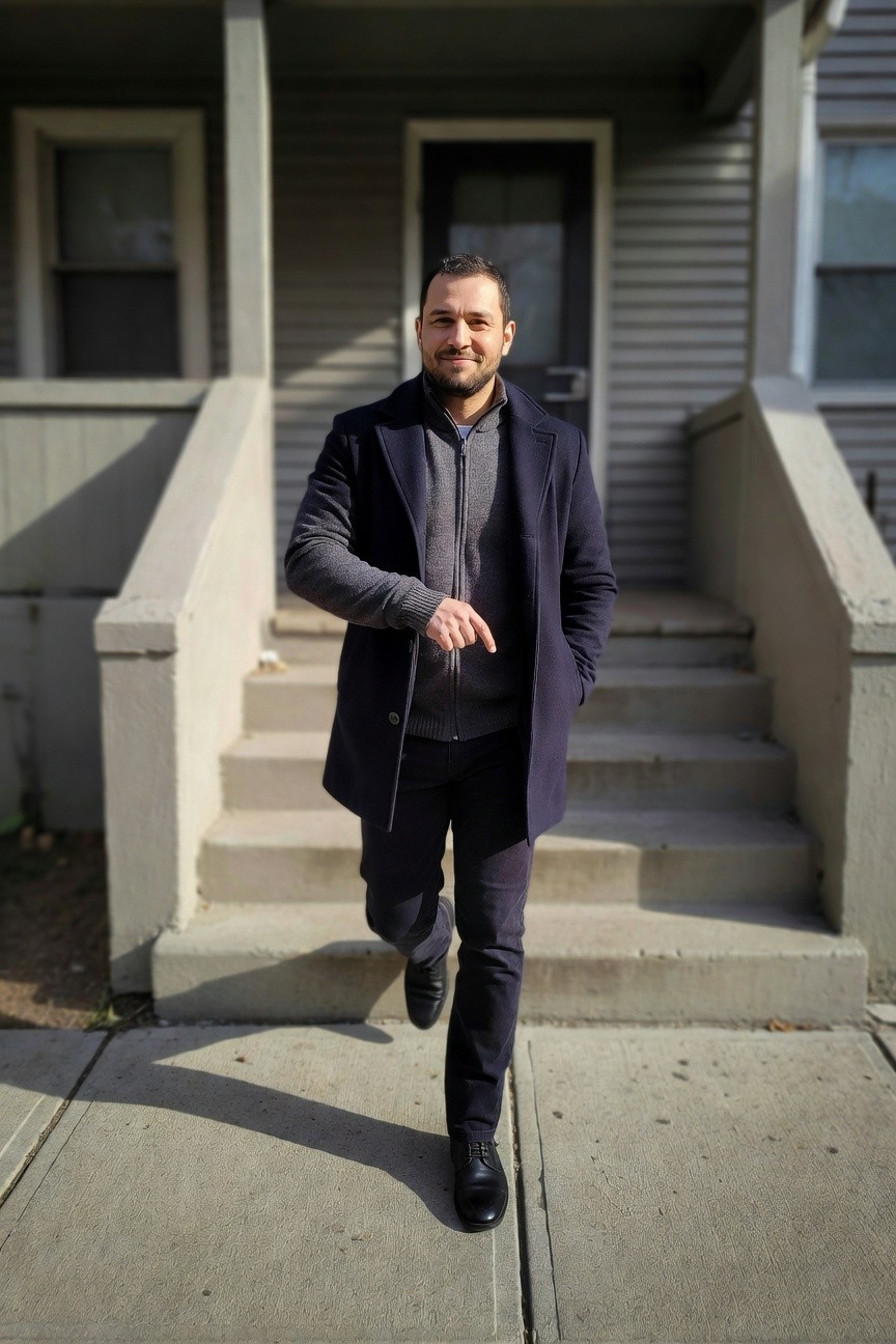 Man in navy wool overcoat over gray zip-up hoodie, slim dark pants, and black leather shoes, standing on concrete steps