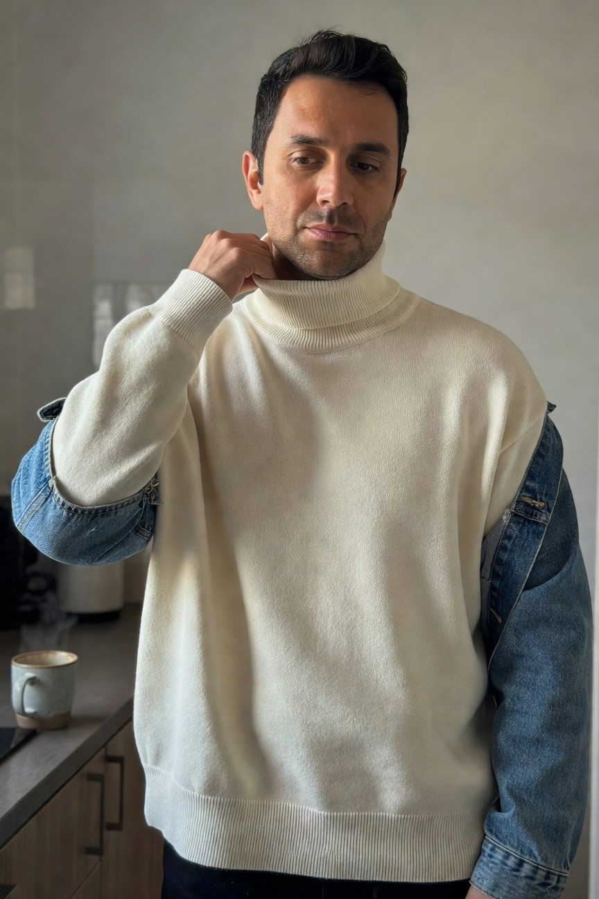 Man in cream turtleneck sweater with integrated blue denim sleeves and shoulders over black pants, hand adjusting collar in indoor kitchen setting