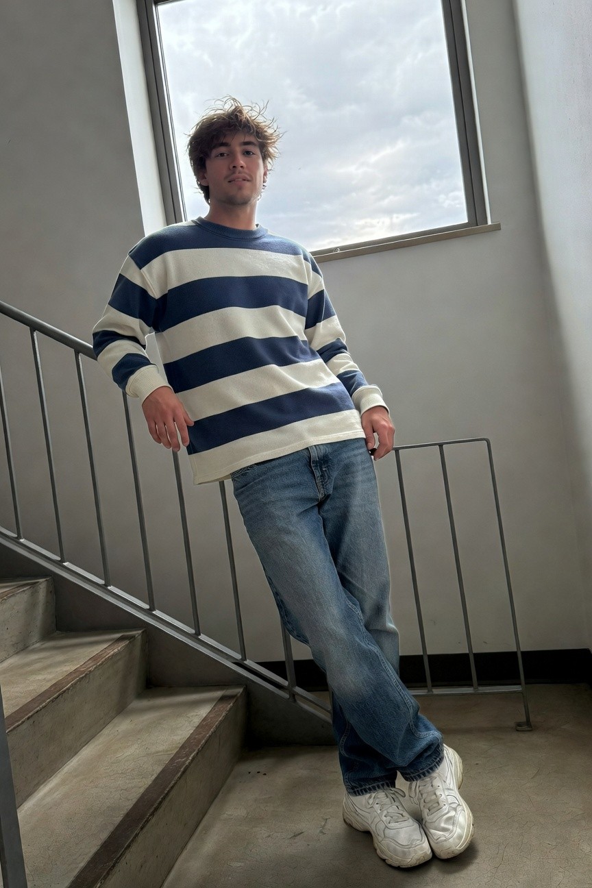 Young man leaning on metal railing wearing navy and white horizontal striped long-sleeve cotton sweater, straight-leg blue jeans, and white chunky sneakers, casual pose on concrete stairs indoors