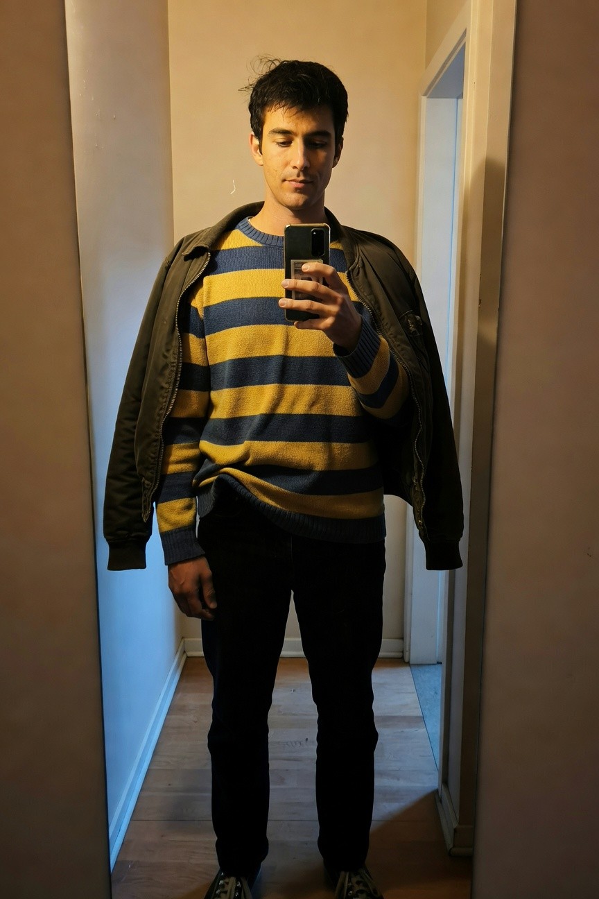 Young man taking mirror selfie in hallway, wearing mustard yellow and navy striped knit sweater layered under open olive green bomber jacket, slim black pants, and black chunky sneakers with metallic accents