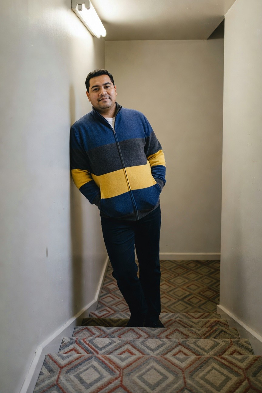 Man leaning against wall in narrow stairwell wearing navy zip-up hoodie with gray stripes and yellow bands on sleeves and chest over white shirt, slim dark trousers, and dark shoes, smiling casually