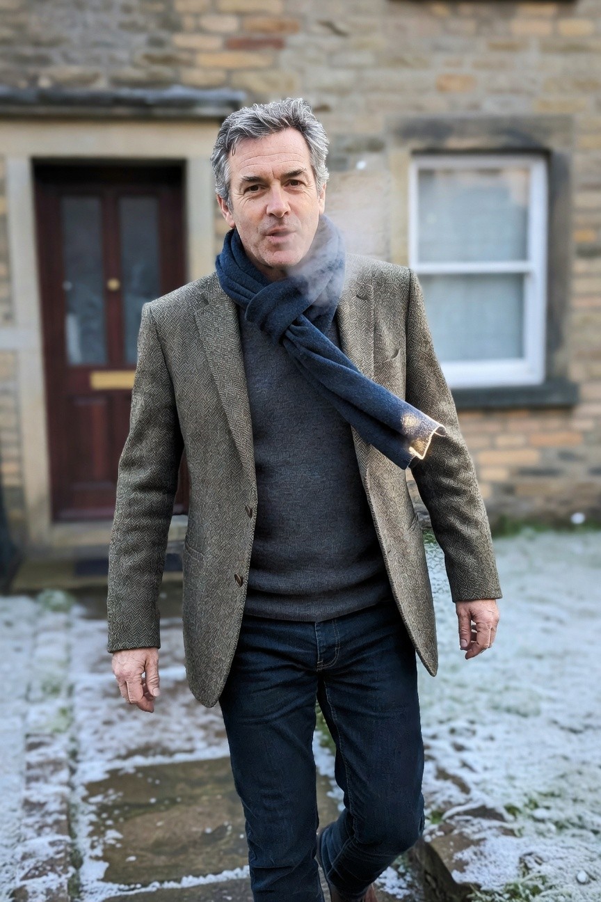 Man in gray herringbone tweed blazer layered over dark gray sweater with navy scarf draped across shoulder, dark blue jeans, and brown boots, standing casually outdoors