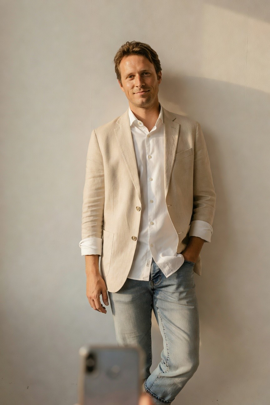 Man leaning against wall in beige linen blazer over white shirt with sleeves rolled, light blue cuffed jeans, holding phone, casual polished style