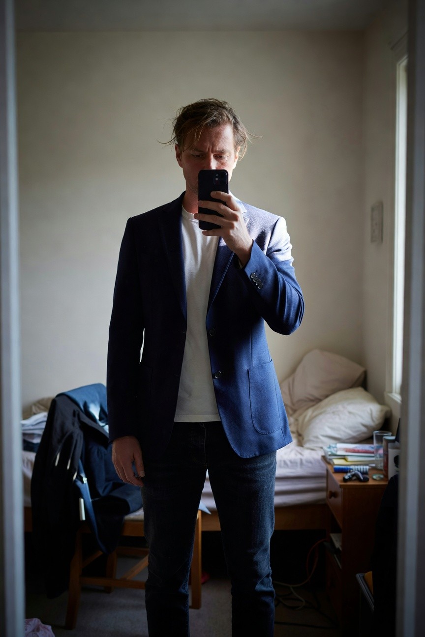 Man in navy blue blazer over white t-shirt and dark slim jeans, taking a mirror selfie in a simple bedroom setting
