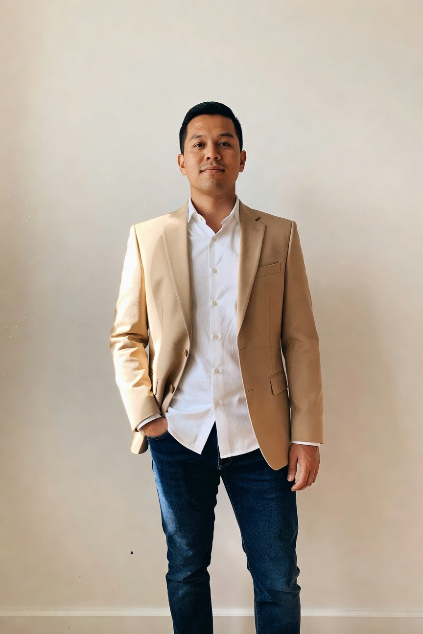Man in light tan slim-fit blazer over white button-up shirt and dark blue slim jeans, hand in pocket, standing against white wall