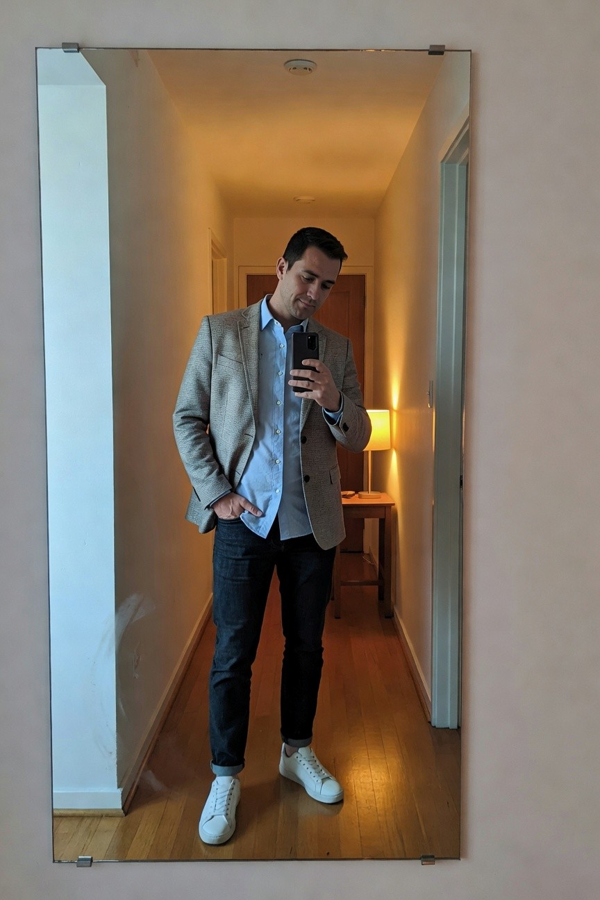 Man in a light gray textured blazer over a light blue button-up shirt, dark slim jeans cuffed at the ankles, and white low-top sneakers, posing casually with hands in pockets in front of a full-length mirror
