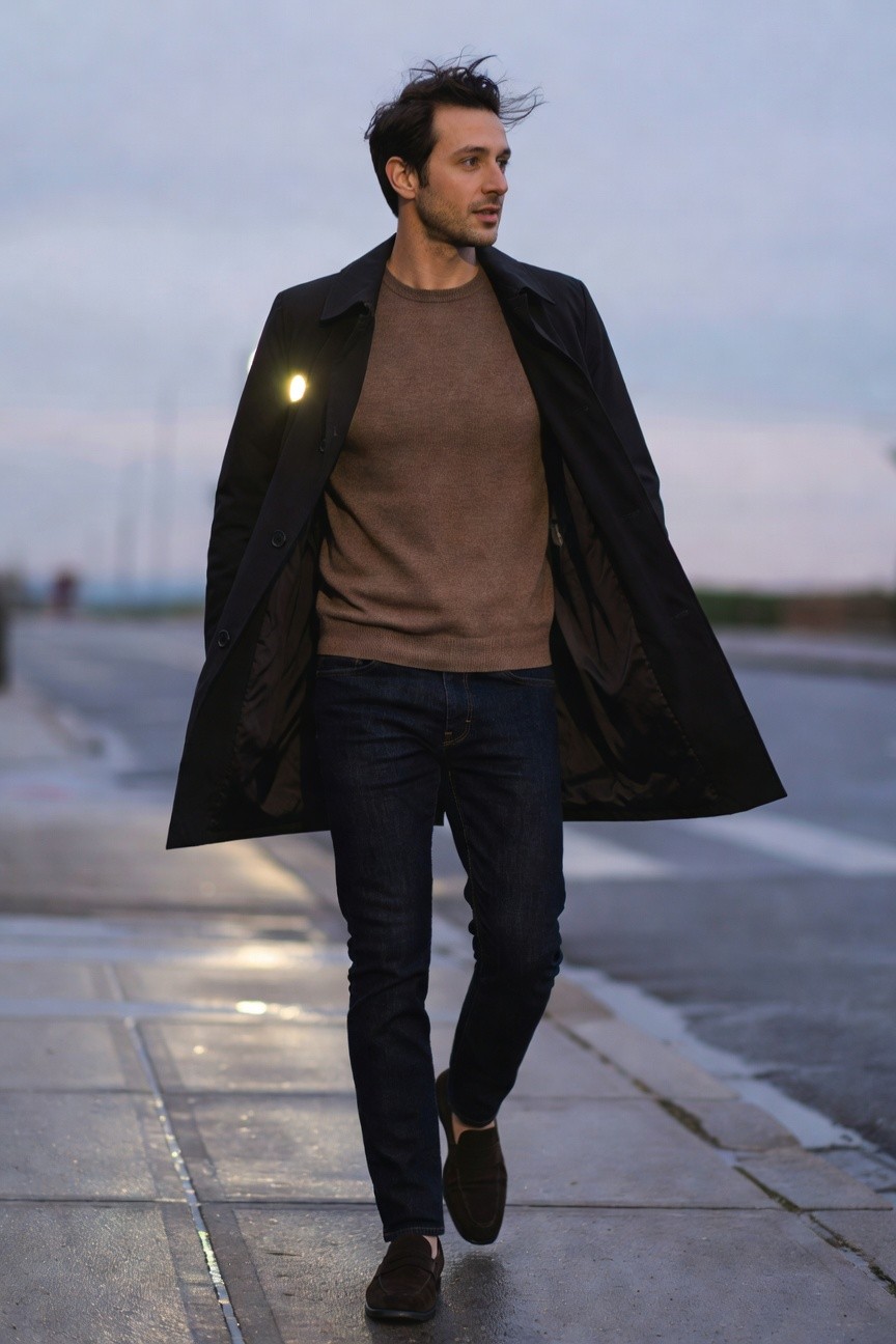 Man walking in black overcoat over beige sweater, dark jeans, and brown loafers on wet pavement