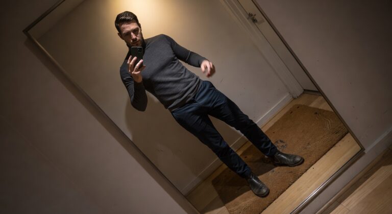Man in dark gray turtleneck sweater, navy chinos, and brown Chelsea boots posing in a dimly lit hallway mirror selfie
