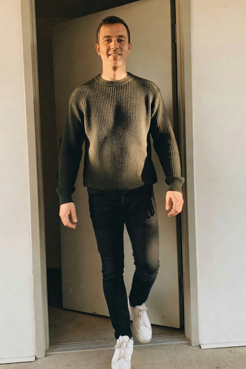 A man stands smiling in an olive green ribbed knit crewneck sweater, black slim-fit jeans, and white sneakers, posed casually in a doorway.