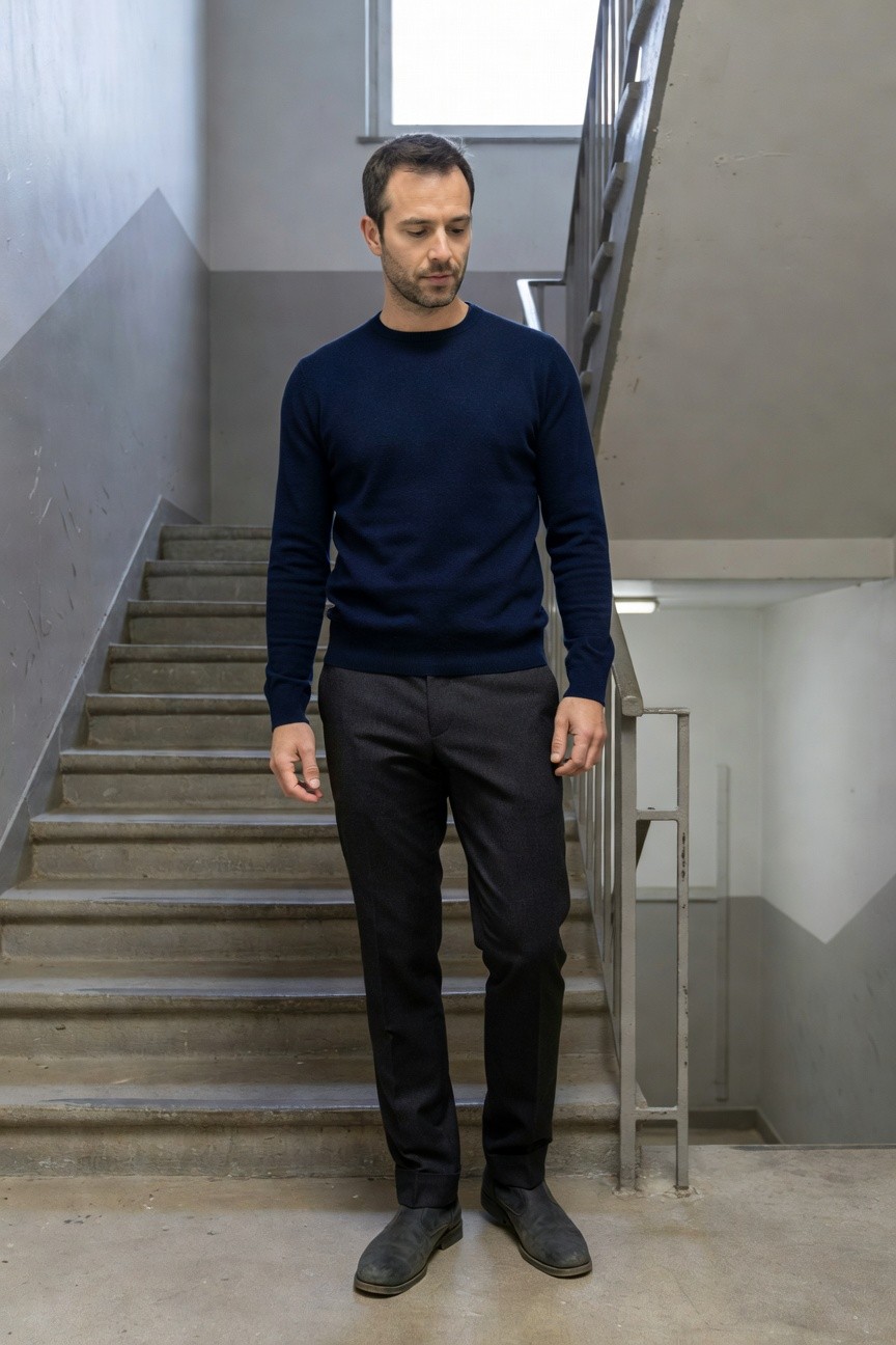 Man in navy crewneck sweater, gray wool trousers, and black Chelsea boots standing on concrete stairs in a modern stairwell, refined evening outfit